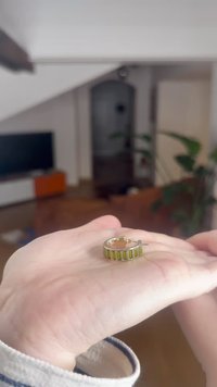 Gullørering med grønne rektangulære steininnlegg. Glatt finish, sirkulært design, vist på en hånd mot en uskarp bakgrunn.