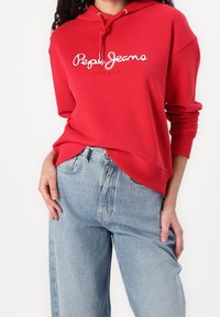 Rode katoenen hoodie met wit logo "Pepe Jeans London," met een losse pasvorm, geribbelde manchetten en een capuchon met trekkoord. Gecombineerd met lichtblauwe spijkerbroek.