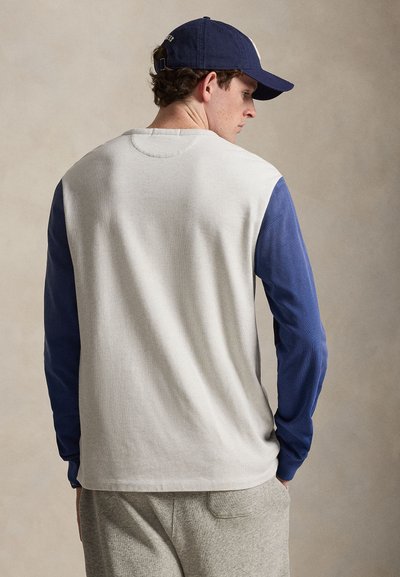 Fiatal férfi, aki fehér inget kék ujjakkal, szürke nadrágot és tengerészkék baseballsapkát visel, háttal áll egy egyszínű háttér előtt.