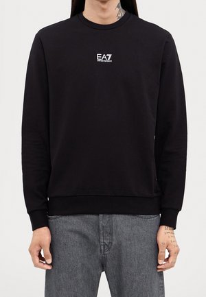 Persoon draagt een zwarte sweatshirt met lange mouwen met een klein wit "EA7 Emporio Armani" logo op de borst en grijze spijkerbroek.