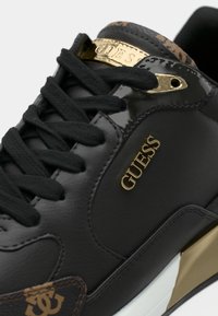 Sorte lærersneakers med gulldetaljer, logo, teksturerte aksenter, svarte snøringer og blanke sidepaneler med et signaturmønster.