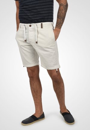 Pantaloni corti beige chiaro in misto cotone con vita regolabile con cordoncino e dettagli marroni. Il modello indossa una camicia a righe scure e scarpe slip-on blu navy.