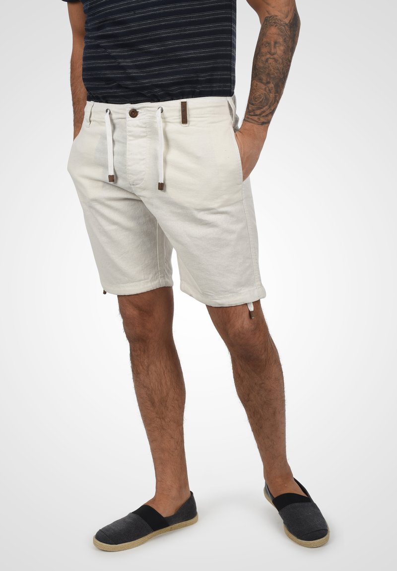 Licht beige shorts van een katoenmix met een trekkoord in de taille en bruine accenten. Het model draagt een donker gestreept shirt en marineblauwe instappers.