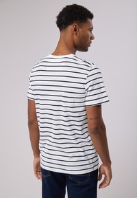 Valkoinen puuvillapaita, jossa on mustia vaakasuoria raitoja, lyhyet hihat, pyöreä kaula-aukko ja suora helma, henkilön päällä näkyy takaa.