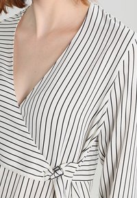 Weißes Wickeloberteil mit schwarzen vertikalen Streifen, ausgestattet mit V-Ausschnitt, langen Ärmeln und einem Metallring-Akzent zum Verschließen an der Taille.