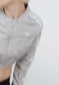 Svetlo šedá športová bunda na zips s tromi bielymi pruhmi na rukávoch a malým logom na ľavej hrudi, v kombinácii s čiernym spodným dielom.