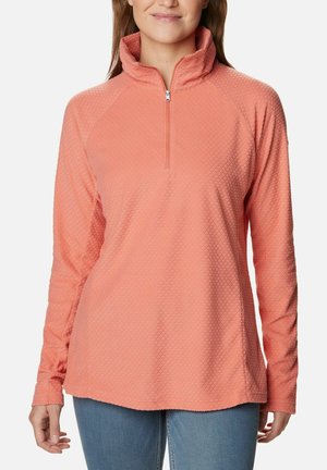 Frau trägt einen strukturierten korallenrosa Langarmpullover mit Viertel-Reißverschluss-Kragen und hellblauen Jeans, steht vor einem schlichten Hintergrund.