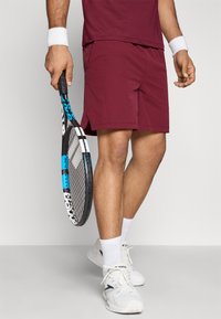 Bordové športové šortky s bočnými rozparkami, v kombinácii s bielymi ponožkami a tenisovými topánkami. Drží tenisovú raketu s čierno-modrým dizajnom.