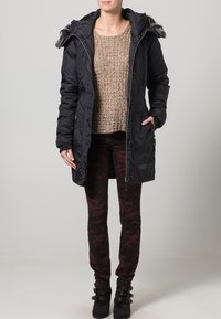 Svart vadderad parkas med huva kantad med päls, dragkedja framtill och sidofickor, kombinerad med en beige tröja och vinröda leggings med mönster.