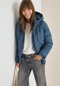 Giacca imbottita blu con cappuccio, caratterizzata da linee di trapuntatura diagonali e cordoncino regolabile. Indossata sopra un maglione crema e jeans grigi.
