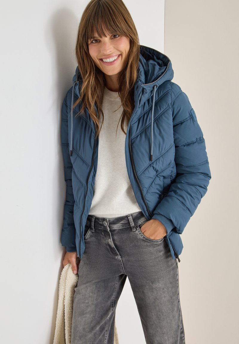Giacca imbottita blu con cappuccio, caratterizzata da linee di trapuntatura diagonali e cordoncino regolabile. Indossata sopra un maglione crema e jeans grigi.