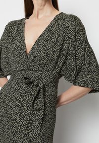 Robe noire avec un motif abstrait vert, décolleté en V profond, manches amples et un lien à la taille. Tissu doux avec une apparence texturée.