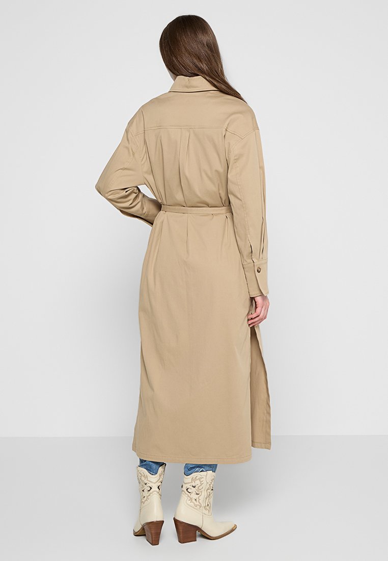Beżowy długi płaszcz z kołnierzykiem, paskiem w talii i bocznymi rozcięciami. Wykonany z gładkiego materiału, zestawiony z jasnymi kowbojkami.