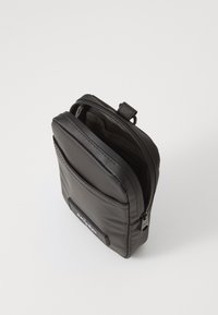 Sac noir en nylon avec un compartiment principal zippé, poche avant et patch logo. Texture lisse et forme rectangulaire.