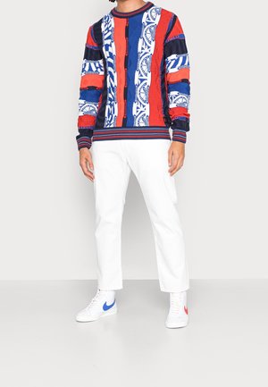 Bunter Strickpullover mit roten, blauen und weißen Mustern; kombiniert mit weißen Hosen und weißen Sneakers mit roten und blauen Akzenten.