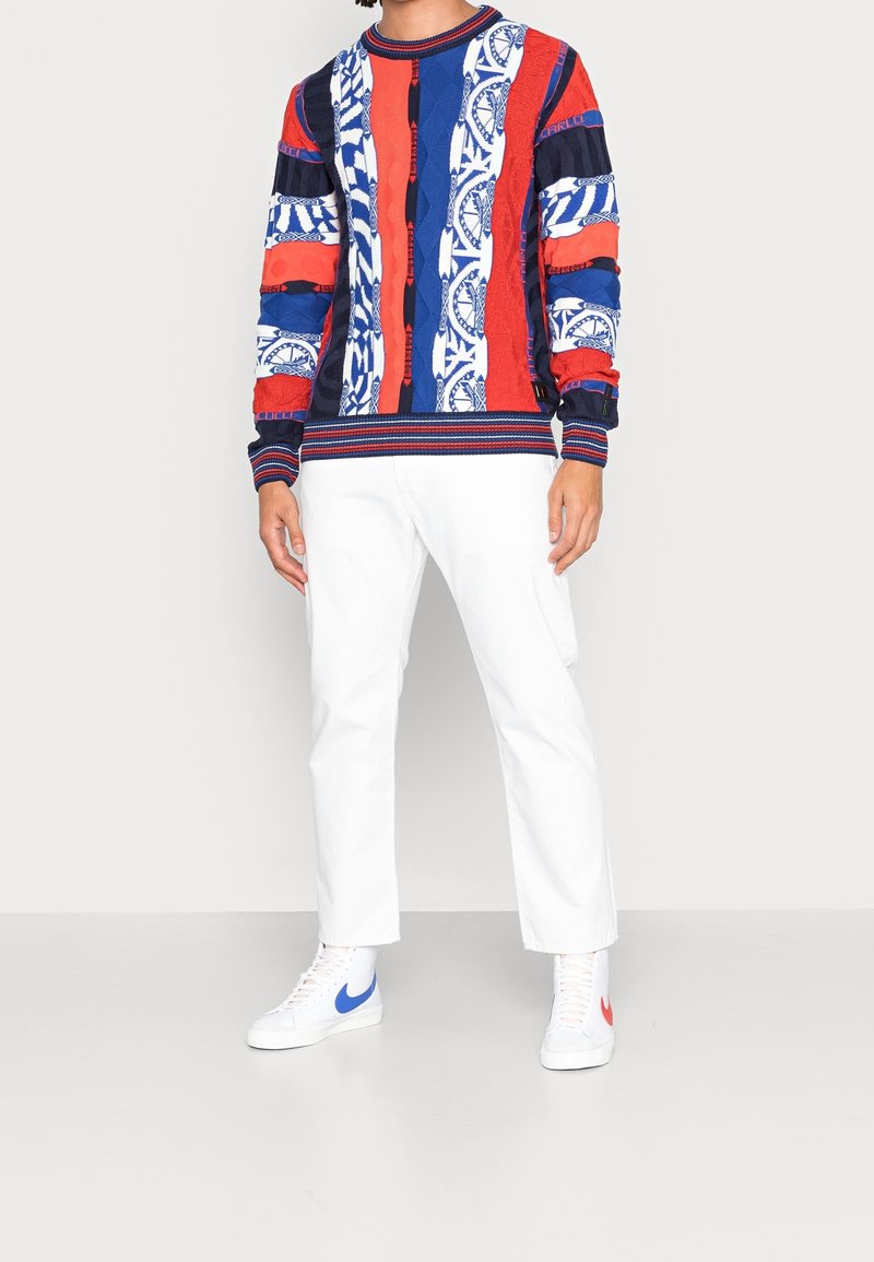 Kleurvolle gebreide trui met rode, blauwe en witte patronen; gecombineerd met witte broeken en witte sneakers met rode en blauwe accenten.