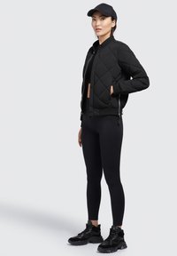 Zwart gewatteerd bomberjack met zijknippers, gecombineerd met strakke zwarte leggings en gestructureerde zwarte sneakers, gedragen door een model.