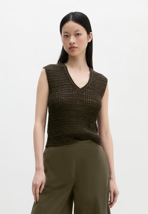 Jeune femme aux cheveux noirs raides portant un haut en crochet marron foncé sans manches et un pantalon taille haute vert olive sur un fond uni.