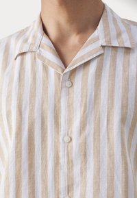 Gros plan d’une chemise beige et blanche à rayures verticales, boutonnée, avec un col ouvert portée par une personne.