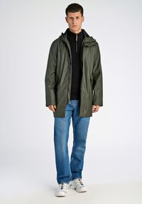 Chaqueta verde impermeable con capucha, que cuenta con cierre de cremallera y botones a presión. Llevada sobre un suéter negro de punto, combinada con jeans azules y zapatillas blancas.