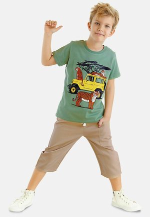 Grünes Baumwoll-T-Shirt mit einem Tiger- und gelben Jeep- Grafik, kombiniert mit beigen, verkürzten Hosen und weißen High-Top Sneakern.