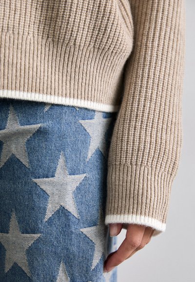 Beige Strickpullover mit gerippter Textur und weißen Akzenten, über blauen Denim mit großem weißen Sternenmuster getragen.