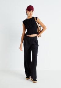 Čierny uzlíkový crop top skombinovaný s čiernymi nohavicami s vysokým pásom. Modelka má na sebe bordovú čiapku a nesie béžovú kabelku cez plece. Čierne topánky s platformou.