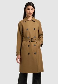 Donna che indossa un trench marrone doppiopetto fino al ginocchio con cintura, in piedi con una mano in tasca contro uno sfondo semplice.