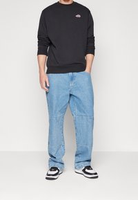 Sudadera negra con cuello redondo, logo rojo. Pantalones de mezclilla holgados en azul claro con detalles de panel. Zapatillas negras y blancas con cordones.
