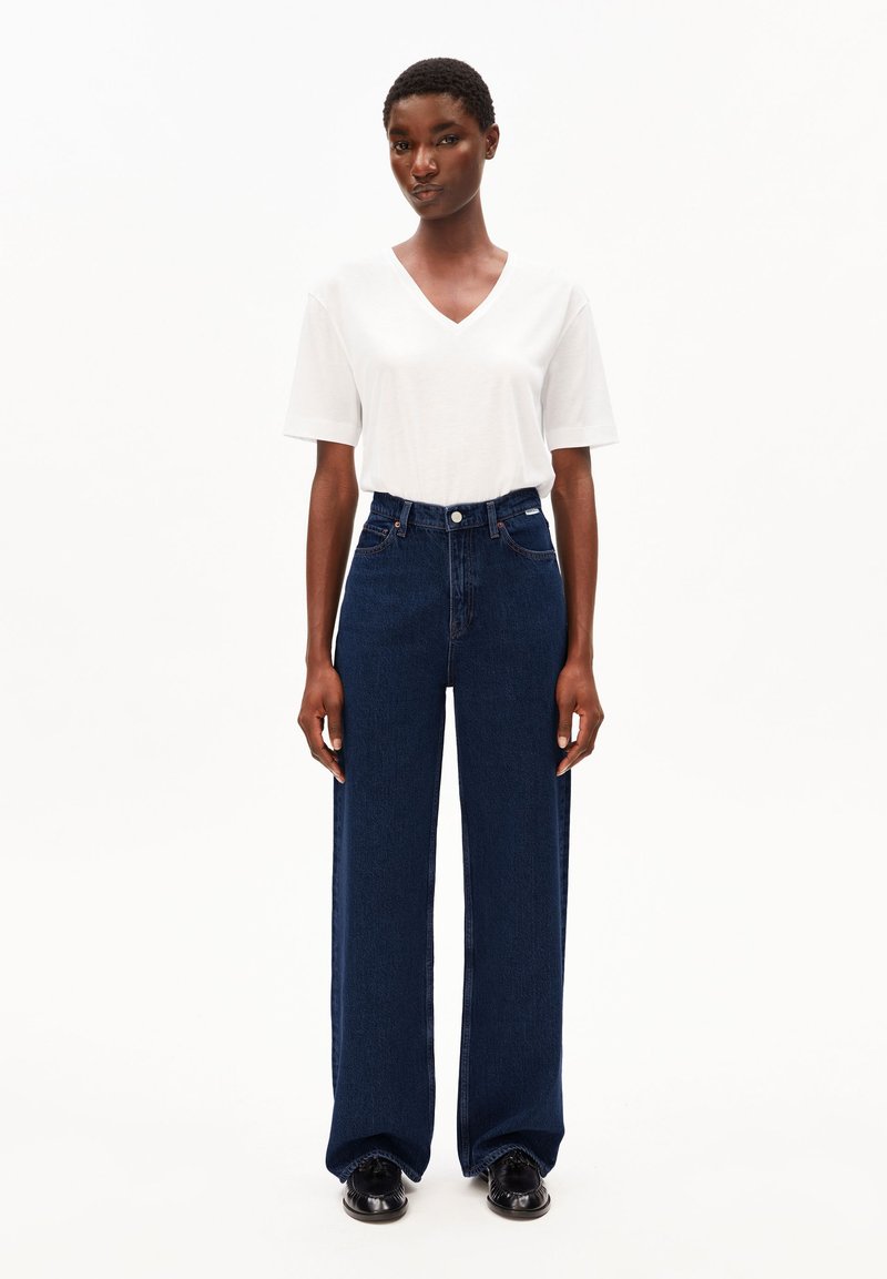 Person steht vor weißem Hintergrund und trägt ein schlichtes weißes V-Ausschnitt-T-Shirt, hoch taillierte dunkelblaue Jeans mit weitem Bein und schwarze Schuhe.