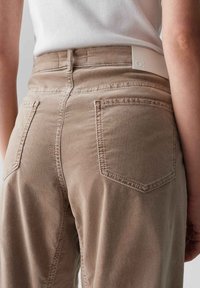 Pantaloni in twill di cotone beige con tasche posteriori, una texture liscia, cuciture visibili e una patch con logo sulla cintura.