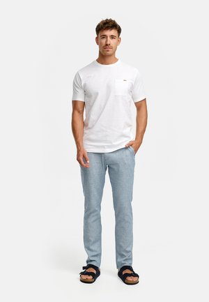 Vit bomulls-t-shirt med bröstficka, ljusblåa texturerade byxor och mörka sandaler med rosett, stående på en enfärgad bakgrund.