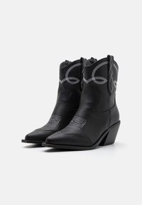 Par črnih usnjenih kavbojskih škornjev z belo okrasno vezenino in poševnimi naloženimi petami na belem ozadju.