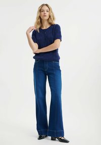 Maglione lavorato a maglia blu navy con maniche corte e motivo testurizzato, abbinato a jeans a zampa d'elefante blu scuro e décolleté nere a punta.