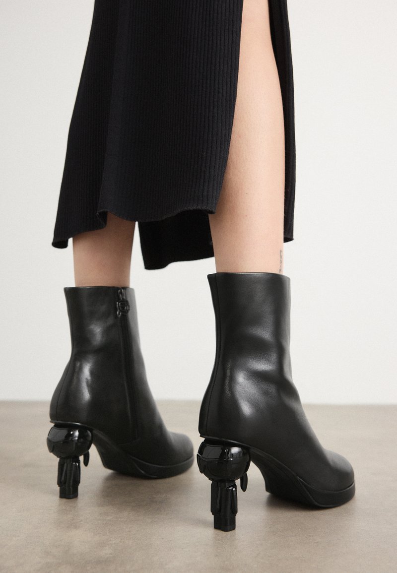 Schwarze Leder-Stiefeletten mit runder Zehenform, die über ein einzigartiges skulpturales Absatzdesign verfügen, das einer kleinen Figur ähnelt. Glatte Textur, Reißverschluss-Detail.