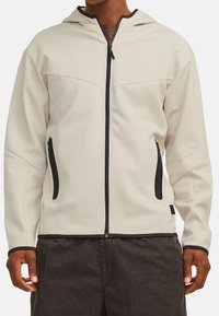 Haut à capuche zippé beige clair avec une capuche, fermeture éclair noire et deux poches latérales. Fabriqué avec un tissu lisse et des accents noirs contrastants.
