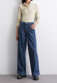 Kobieta w beżowej bluzie z zamkiem błyskawicznym i długim rękawem, w spodniach z szeroką nogawką w niebieskim kolorze z wysokim stanem oraz czarnych butach na grubych podeszwach, stojąca na tle białej ściany.