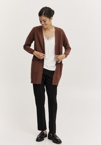 Cardigan tricotat de culoare maro, cu fața deschisă, buzunare laterale și mâneci lungi, stratificat peste un top alb cu dantelă în jurul gâtului și pantaloni negri.