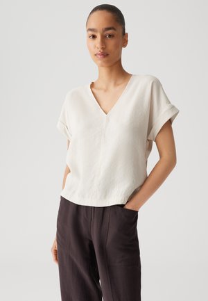 Crème-kleurige blouse met korte mouwen, een V-hals en een ruwe rand, gecombineerd met donkere broek met zijzakken en een relaxed fit.