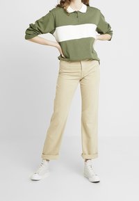 Personne debout les mains sur les hanches, portant un t-shirt à manches longues rayé vert olive et blanc, un pantalon beige retroussé aux chevilles et des baskets blanches.