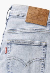 Jeans in denim azzurro chiaro presentano una patta in pelle con logo impresso, cuciture a contrasto e una caratteristica etichetta rossa sulla tasca posteriore.