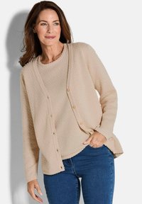 Beige Strickjacke mit einem strukturierten Muster, rundem Halsausschnitt und Knopfverschluss. Lange Ärmel und eine lockere Passform. Getragen über dunkelblauen Jeans.