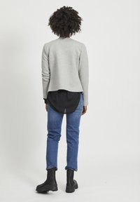 Gray textured jacket with a rounded hem, worn over a longer black shirt, paired with blue jeans and black ankle boots. Rear view displayed.