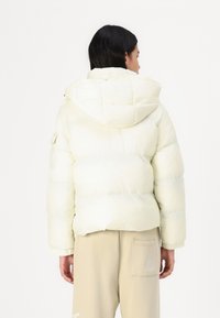 Dikke, lichtgele jas met een capuchon, met gestikte delen en een gladde textuur. Gecombineerd met beige broek, met een detail van een zak.