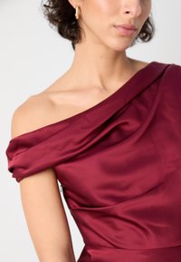 Bordo satino suknelė su pečių neskleidžiančiu iškirpimu, drapiruota medžiaga ir lygi tekstūra. Turinti prigludusią liemenę, pabrėžia eleganciją.