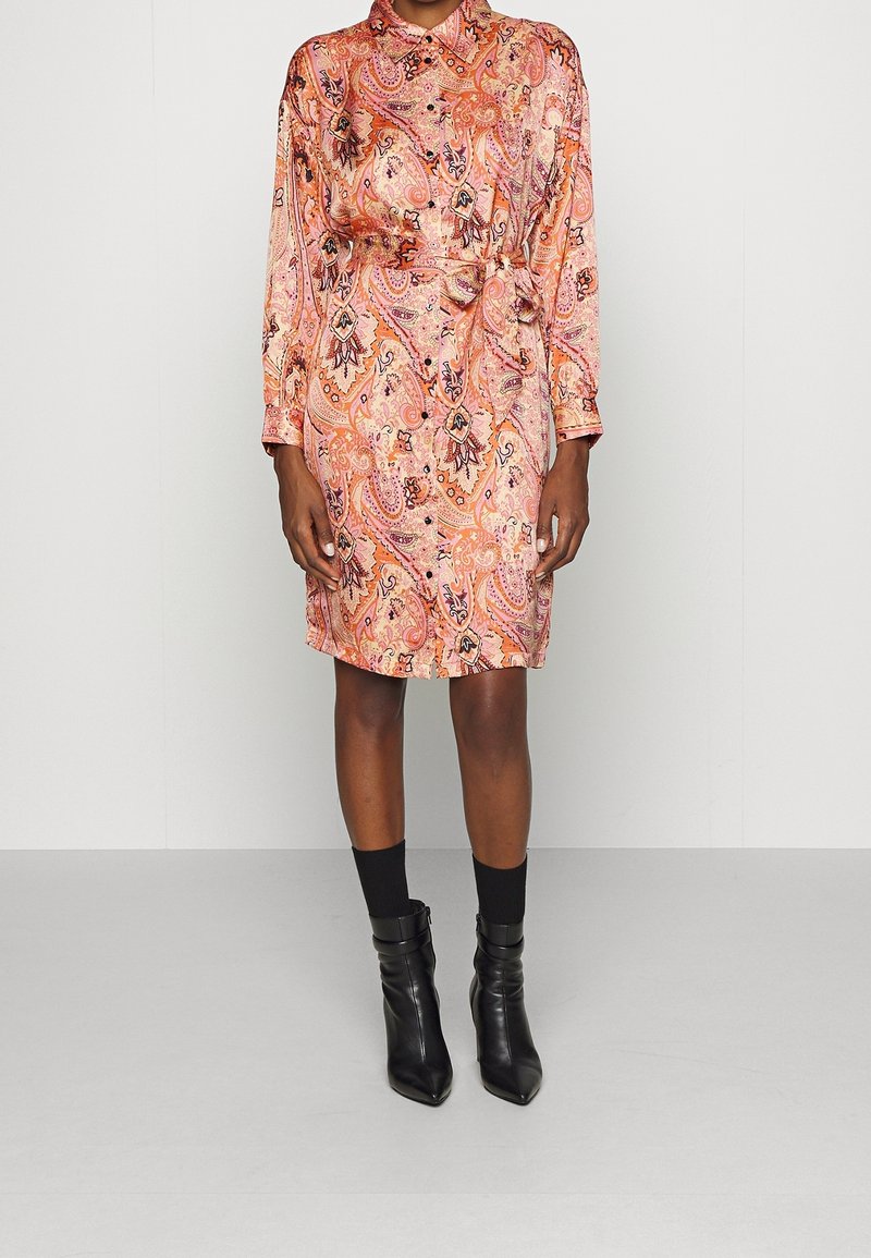 Personne portant une robe à manches longues, longueur genoux, à motif cachemire dans des tons roses et orange, associée à des chaussettes noires et des bottines.