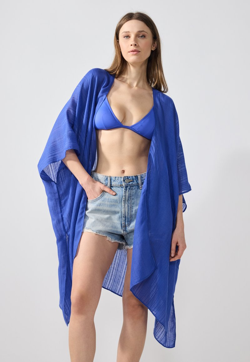 Blå, lätt kimono-liknande överdrag med öppen front, texturerad tyg och vida ärmar, som bärs över en blå bikini och denimshorts.