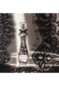 Flacon de parfum élégant portant l'étiquette Christina Aguilera, décoré de dentelle noire et d'un petit nœud, entouré de tissu en dentelle noire.