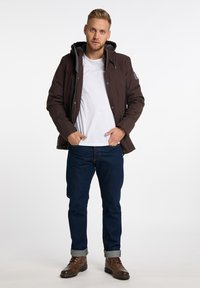 Giacca cappuccio marrone scuro con zip e chiusura a scatto, indossata sopra una maglietta bianca. Abbinata a jeans blu scuro e stivali marroni.