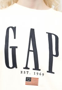 Haut blanc avec de grandes lettres noires "GAP", texte "EST. 1969" en dessous, et un petit drapeau colonial américain brodé.